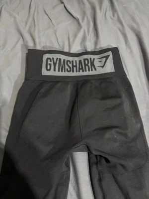 Svarta leggings från Gymshark - Säljer ett par svarta leggings från Gymshark med hög midja och bred resår med logga framtill. Tight passform och stretchigt material som passar perfekt till träning eller chill. Klassisk och stilren design med diskret struktur i tyget.