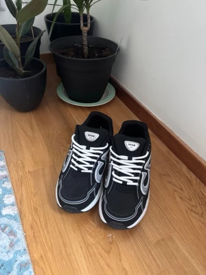 Svarta Dior sneakers med vita detaljer - Snygga svarta sneakers från Dior med vita och grå detaljer samt stora CD-loggor på sidorna. Skorna har vita snören och en chunky vit sula. Ovandelen är i mesh och syntetmaterial för en modern look. Perfekta för dig som vill sticka ut med exklusiva sneakers.