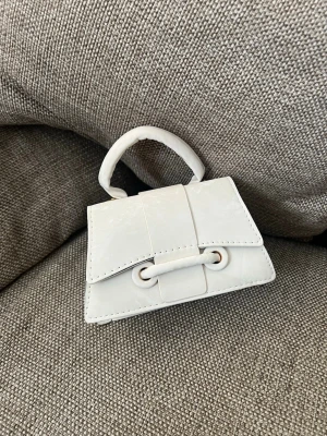 Vit minihandväska med axelrem - Superfin liten vit handväska i blankt material med både handtag och justerbar axelrem. Väskan har en unik design med dekorativ knutdetalj framtill och guldfärgade metalldetaljer. Perfekt för dig som gillar trendiga accessoarer och vill sticka ut.