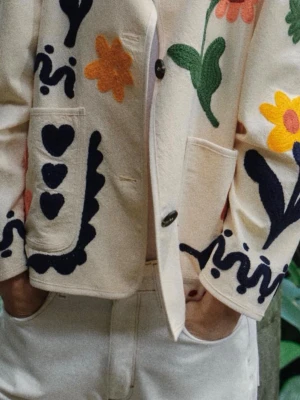 Beige kavaj med broderade blommor - Unik beige kavaj med stora broderade blommor i rosa, gult och grönt samt svarta grafiska detaljer. Rak passform, två stora fickor framtill och knäppning med svarta knappar. Materialet ser ut att vara bomull eller linne, perfekt för dig som gillar färg och kreativ stil.