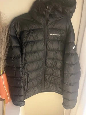 Svart dunjacka Peak Performance - Snygg svart dunjacka från Peak Performance med huva och dragkedja framtill. Jackan har diskret logga på bröstet och är quiltad med breda kanaler. Perfekt för kalla vinterdagar och riktigt skön att bära.