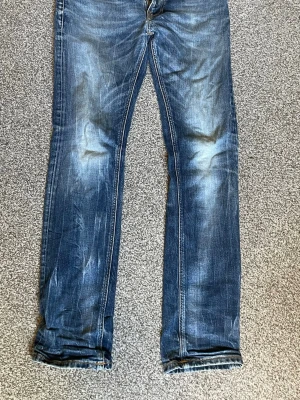 Blå nudie jeans med slitningar - Säljer ett par blå jeans med raka ben och snygga slitningar framtill och baktill. Jeansen har klassisk femficksdesign och kontrastsömmar. Perfekta för en avslappnad och trendig look. Det är ett par nudie jeans som är storleken: W:30 och L:32, öppen för diskutering om pris! 