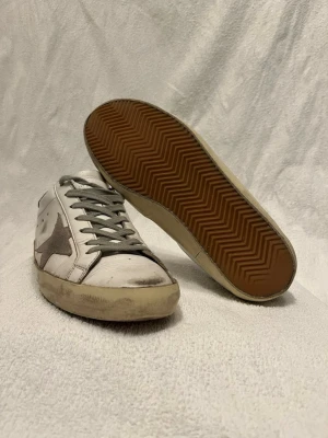 Golden goose skor - Snygga vita sneakers från Golden Goose med grå stjärna på sidan och gråa skosnören. Skorna är i bra skick enda defekten är att dem har blivit proffsiotinellt lagade på sidan, storlek 41 men mer som 41,5 passar dig som har 42 nypris 5000 tusen!!!
