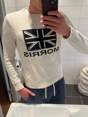 Vit stickad tröja från Morris - Vit stickad tröja från Morris med mörkblå Union Jack-flagga och logga framtill. Tröjan har rund halsringning, ribbade muddar och är långärmad. Perfekt för en stilren och klassisk look. Modellen är 187 cm och väger 80 kg