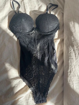 Svart spetsbody från H&M - Säljer en svart body i spets med djup v-ringning och tunna axelband. Bodyn har transparenta partier på sidorna och ett detaljerat spetsmönster framtill. Perfekt för dig som gillar en stilren och lite sexigare look.