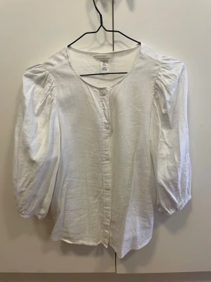 Vit linnemixblus från H&M - Snygg vit blus från H&M i linnemix med puffiga trekvartsärmar och rund halsringning. Blusen har knäppning framtill och en avslappnad passform som ger en trendig och luftig känsla. Perfekt att styla med jeans eller kjol.