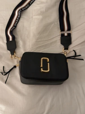 Marc Jacobs Snapshot - Snygg svart axelväska från Marc Jacobs med guldiga detaljer och bred, randig axelrem. Väskan har dubbla dragkedjor och präglad logga på sidan. Tillverkad i strukturerat läder och har en stilren, boxig form. Den är lite smutsig runt om logon.