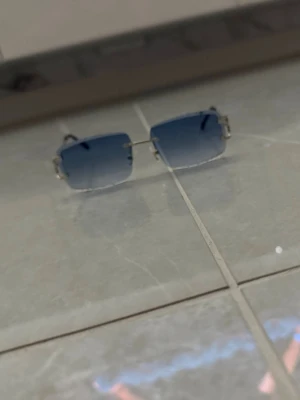 Blå rektangulära solglasögon - Snygga solglasögon med rektangulära blåtonade glas och tunna silvriga bågar. Glasen är utan ram och har en modern vibe som passar perfekt till sommaren. Enkla att matcha med olika outfits och ger en cool touch till din stil.