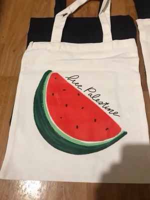 Vit tygkasse med vattenmelontryck - Snygg vit shoppingväska i tyg med ett stort tryck av en vattenmelon och texten 'Free Palestine' på framsidan. Perfekt att bära böcker, mat eller annat i. Väskan har långa handtag och är lätt att vika ihop när den inte används.