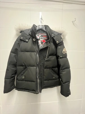 Svart dunjacka från Moose Knuckles - Snygg svart dunjacka från Moose Knuckles med avtagbar huva och lyxig pälsdetalj. Jackan har flera fickor med dragkedja och tryckknappar samt den ikoniska logotypen i metall på ärmen. Perfekt för kalla dagar och riktigt varm.