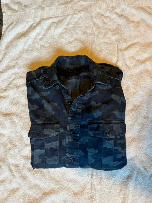 Ralph Lauren skjorta - Snygg mörkblå skjorta med camouflage-mönster från Polo Ralph Lauren. Skjortan har två bröstfickor med knappar och klassisk krage. Perfekt för dig som vill ha en cool och avslappnad stil.