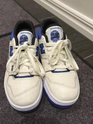 New Balance 550 vita/blå sneakers - Säljer ett par New Balance 550 sneakers i vitt och blått med svarta detaljer. Skorna har klassisk låg siluett, perforerad tå och tydlig New Balance-logga på sidan. Ovandelen är i läder och mesh för en sportig look.