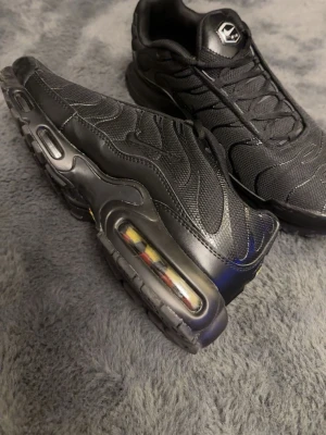 Nike Air Max Plus svarta sneakers - Nike Air Max Plus sneakers i helsvart med mesh och syntetiska paneler. Snygga vågformade detaljer på ovandelen och synlig Air Max-dämpning i sulan. Klassisk logga på plösen och robust yttersula med bra grepp. Perfekta för dig som gillar streetwear och sportig stil.
