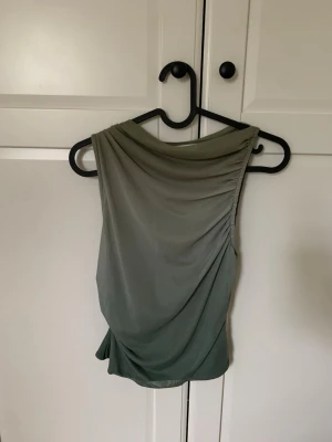 Grön topp från Pull&Bear - Snygg ärmlös topp från Pull&Bear i olika gröna nyanser med draperad effekt. Toppen har en mjuk, följsam passform och är perfekt för att skapa en trendig look. Tillverkad i ett lätt och stretchigt syntetmaterial.