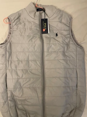 Ljusgrå dunväst från Polo Ralph Lauren - Ljusgrå dunväst från Polo Ralph Lauren med klassisk ståkrage och dragkedja framtill. Västen har diskret broderad logga på bröstet och quiltad design. Perfekt för lager-på-lager och snygg till både hoodie och t-shirt.