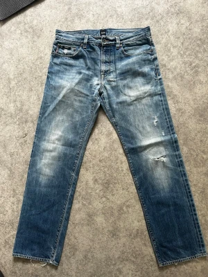 Blå slitna jeans från Hugo boss - Säljer ett par blå jeans från Hugo boss  med raka ben, slitningar och trasiga detaljer på framsidan. Jeansen har klassisk femficksdesign, ljusa tvättade partier och råa kanter vid bensluten. Perfekta för en avslappnad streetstil.