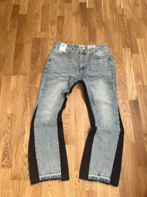 Jeans Gallery Dept. La Flare Two-Tone  - These GALLERY DEPT. jeans feature a light wash denim with striking black panels along the outer seams and stylish raw, distressed hems. The distinctive two-tone design and flared cut embody the brand's unique artistic vision and streetwear appeal, making them a standout piece.  