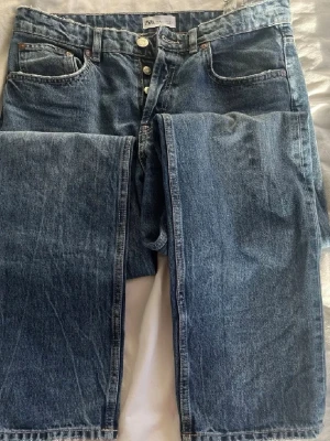 Blå raka jeans från Zara - Snygga blå jeans från Zara med rak passform och klassisk femficksdesign. Jeansen har knäppning med knappar och är gjorda i denim med en lätt tvättad look. Perfekta för dig som gillar en avslappnad och tidlös stil.