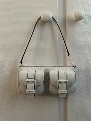 Vit axelremsväska Michael Kors - Snygg vit axelremsväska från Michael Kors med två framfickor och silverfärgade spännen. Väskan har MK-logga i mönstret och detaljer i vitt konstläder. Praktisk och trendig modell med svart kant på remmen och lock på fickorna.