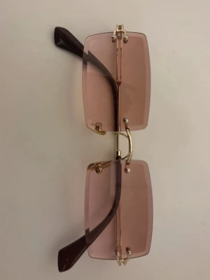 Rosa rektangulära solglasögon - Snygga solglasögon med rektangulära rosa glas och guldfärgade detaljer. Glasögonen har en modern, kantig form och tunna skalmar i mörkbrunt. Perfekta för att lyfta din stil med en trendig touch.