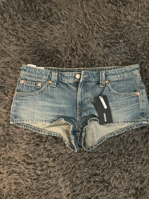 Blå denimshorts från Weekday - Säljer ett par helt oanvända klassiska blå denimshorts från Weekday med fem fickor, bälteshällor och knappgylf. Shortsen har en lågmidjad passform och är tillverkade i bomull. Perfekta för sommaren och ger en avslappnad vibe.