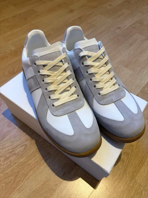 Maison Margiela skor - Maison Margiela sneakers i vitt och ljusgrått läder och mocka. Klassisk låg modell med vita snören och gummisula. Snygg minimalistisk design som passar till det mesta. Märkets ikoniska etikett syns på insidan av skon. Skorna är oanvända och låda ingår. Skriv vid funderingar eller frågor.