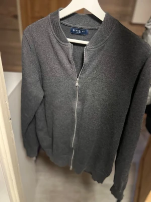 Grå cardigan från Berkeley - Mörkgrå cardigan från Berkeley med rund halsringning och hel dragkedja framtill. Tröjan har långa ärmar och ribbade muddar vid ärmslut och nederkant. Perfekt för en avslappnad och stilren look. Väldigt bra skick inge tecken på användning.