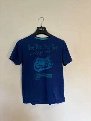 Blå t-shirt med motorcykeltryck - Blå t-shirt med coolt motorcykeltryck och texten 'See That You Get the genuine' framtill. Klassisk passform med rund hals och korta ärmar. Perfekt för dig som gillar avslappnad stil och grafiska tryck.