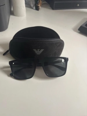 Svarta solglasögon Emporio Armani - Snygga svarta solglasögon från Emporio Armani med klassisk rektangulär form. Glasögonen har svarta bågar och mörka glas, och kommer med ett svart fodral med logga. Perfekta för att lyfta din stil under soliga dagar.