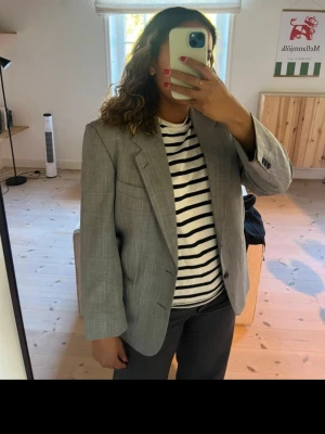 Grå rutig oversized kavaj - Snygg grå kavaj med diskret rutmönster och oversized passform. Klassisk design med två knappar framtill, breda slag och tre fickor. Perfekt för en trendig och avslappnad look. Materialet känns mjukt och har en lätt struktur.