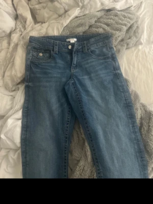 Blå raka jeans med fickdetaljer - Snygga blå jeans med rak passform och klassiska fem fickor. Jeansen har en ljus tvätt och silverfärgade knappar på fickorna. Perfekta till en avslappnad stil och funkar året runt. Materialet är denim i bomull.
