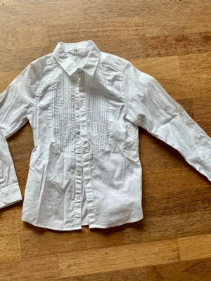 Vit skjorta med veckad front 134 - Klassisk vit skjorta med lång ärm och knappar framtill. Skjortan har snygga veckade detaljer på bröstet och en klassisk krage. Perfekt för en stilren look och enkel att matcha med olika outfits.