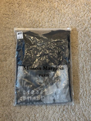 Maison Margiela svart t-shirt - Svart t-shirt från Maison Margiela med tryck framtill i vitt och grått. Motiv med siffror och porträtt samt Maison Margiela Paris-logga. Klassisk rund hals och korta ärmar. Perfekt för dig som gillar streetstyle och designerplagg.