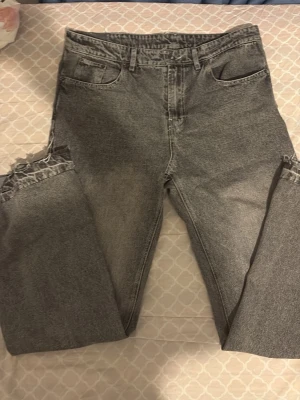 Grå jeans med flared fit - Säljer ett par grå jeans med raka ben och råa, fransiga benslut. Jeansen har klassisk femficksdesign och bälteshällor. Perfekta för dig som gillar en avslappnad och trendig look.