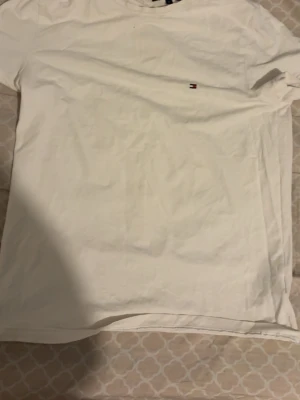 Vit t-shirt från Tommy Hilfiger - Klassisk vit t-shirt från Tommy Hilfiger med liten broderad logga på bröstet. T-shirten har rund hals och korta ärmar. Tillverkad i mjuk bomull som känns skön mot huden. Perfekt basplagg för en clean och stilren look.