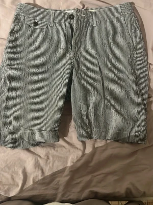Randiga shorts från Helly Hansen - Snygga randiga shorts från Helly Hansen i vitt och mörkblått. Klassisk passform med fickor fram och bak, stängs med knapp och dragkedja. Tillverkade i bomull, perfekta för varma dagar.