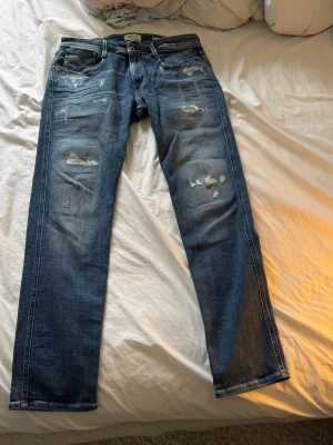 Blå slitna jeans med raka ben från Replay - Säljer ett par blå jeans med raka ben och slitna detaljer på både fram- och baksida från Replay. Jeansen har klassisk femficksmodell och en snygg tvättad look. Perfekta för dig som gillar en avslappnad och trendig stil.