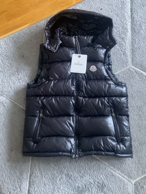 Svart dunväst från Moncler med huva - Snygg svart dunväst från Moncler med glansig finish, avtagbar huva och två dragkedjefickor framtill. Västen har klassisk Moncler-logga på bröstet och quiltad design som ger en modern look. Perfekt för lager-på-lager-stil under kyliga dagar.