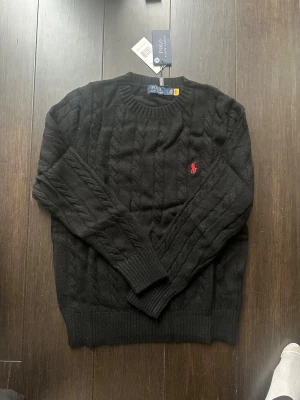 Svart kabelstickad tröja Polo Ralph Lauren - Svart kabelstickad tröja från Polo Ralph Lauren med rund halsringning och röd broderad logga på bröstet. Tröjan har ribbade muddar vid ärmslut och nederkant. Perfekt för en stilren och klassisk look.