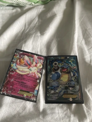 Pokémon EX-kort Sylveon & Blastoise - Två Pokémon EX-samlarspelkort: ett Sylveon EX med rosa och vita färger samt ett Blastoise EX i blått och svart. Båda korten har holografisk finish och är tillverkade i laminerad papp. Perfekt för dig som gillar Pokémon TCG och vill utöka din samling.