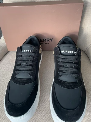 Burberry svarta sneakers med rutigt mönster - Snygga svarta sneakers från Burberry med klassiskt rutigt tyg på hälen, svarta mockadetaljer och vita sulor. Skorna har svarta skosnören och Burberry-logga på plösen. Perfekta för dig som vill ha en stilren och lyxig look. Orörda helt nya med påse i sko kartong. 