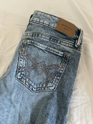 Blå jeans Gina Tricot  - Snygga blå jeans från Gina Tricot. Jeansen har dekorativa sömmar på bakfickan och en brun patch med logga i midjan. Tillverkade i denim med en cool tvättad look.
