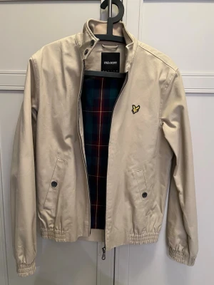 Beige jacka från Lyle & Scott - Stilren beige jacka från Lyle & Scott med klassisk gul logga på bröstet. Jackan har ståkrage, dragkedja, knappar vid fickorna och elastiska muddar. Insidan har rutigt foder i blått och rött. Perfekt för vår och höst.