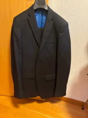 Svart kostym med kavaj och byxor - Snygg svart kostym med klassisk kavaj och matchande kostymbyxor. Kavajen har smala slag, två fickor med lock och en bröstficka. Byxorna har raka ben och bälteshällor. Perfekt för dig som vill ha en stilren och tidlös look. Strlk S