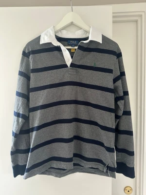  Polo Ralph Lauren strl xs (14-16 år) Randig rugbytröja - Klassisk långärmad rugbytröja från Polo Ralph Lauren i grått med mörkblå ränder och vit krage. Tröjan har knappar vid halsen och den ikoniska gröna loggan broderad på bröstet. Tillverkad i mjuk bomull för en bekväm känsla. Strl 14-16 år motsvarar XS. Mått bröst 42 cm, axlar 42 cm, längd 62 cm. 