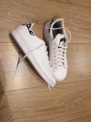 Adidas Stan Smith  - Snygga vita Adidas Stan Smith sneakers i klassisk låg modell. Skorna har vita snören, perforerade ränder på sidorna och svart detalj med vit logga på hälen. Tillverkade i slätt läder för en clean och tidlös look. Kommer i orginal kartong. 