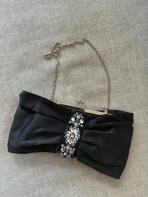 Svart kuvertväska med strassdetaljer - Stilren svart kuvertväska i satin med dekorativ strass och pärlor framtill. Väskan har en silverfärgad kedja och metallspänne upptill. Perfekt accessoar för att lyfta din outfit med lite extra bling.