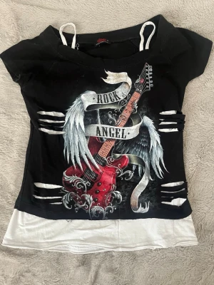 Rock Angel t-shirt med gitarrtryck - Svart t-shirt med tryck av röd elgitarr, vingar och texten 'Rock Angel'. Snygga slitsar i sidorna och vita axelband samt vit nederdel. Perfekt för dig som gillar rockig stil och detaljer.