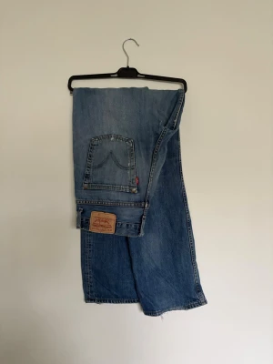 Levi's blå slitna raka jeans - Ett par klassiska blå Levi's jeans med raka ben och slitningar vid knäna. Jeansen har fem fickor, synlig läderpatch bak och den ikoniska röda Levi's-taggen. Tillverkade i kraftig denim, perfekt för en avslappnad streetstil. Storlek 33/35 