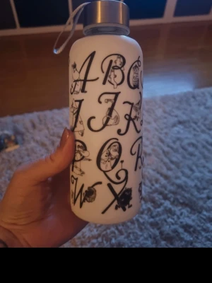 Vit vattenflaska med alfabet-tryck - Säljer en vit vattenflaska i glas och gummi överdrag med svart alfabet-tryck och olika illustrationer vid varje bokstav med mumin. Flaskan har ett silverfärgat skruvlock med bärrem.